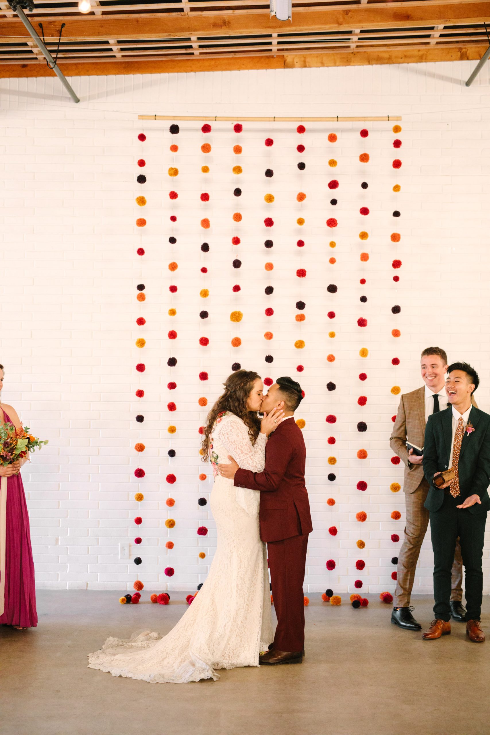 Bride and groom first kiss at wedding ceremony with pom pom backdrop at BLOC Venue Roseville - www.marycostaweddings.com