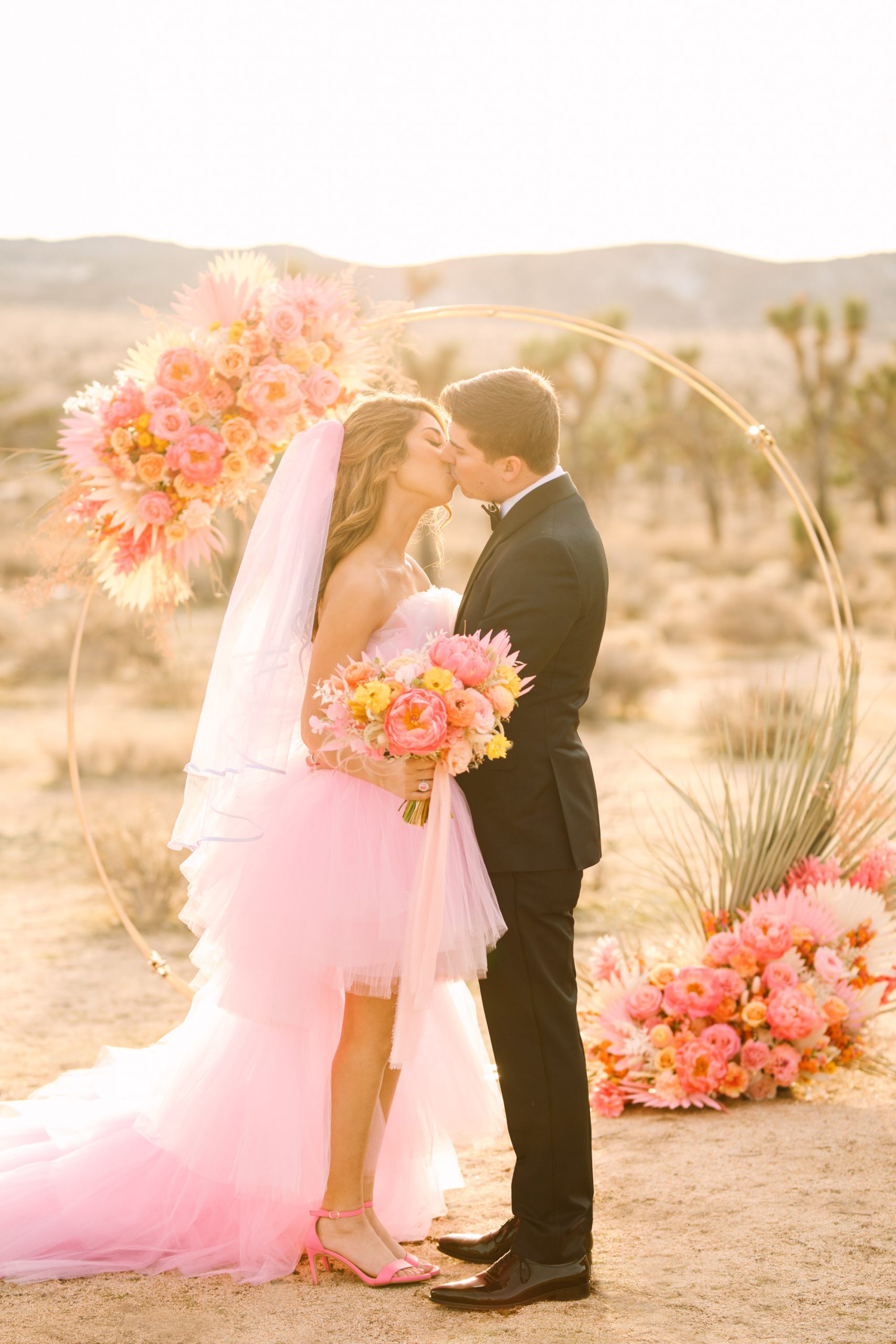 Bride and groom kissing in front of pink floral arch | Pink wedding dress Joshua Tree elopement featured on Green Wedding Shoes | Colorful desert wedding inspiration for fun-loving couples in Southern California #joshuatreewedding #joshuatreeelopement #pinkwedding #greenweddingshoes Source: Mary Costa Photography | Los Angeles
