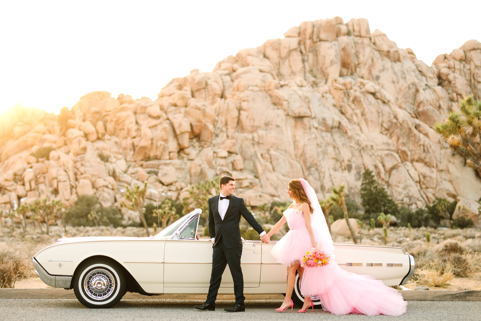 Bride and groom walking with white classic Ford Thunderbird convertible | Pink wedding dress Joshua Tree elopement featured on Green Wedding Shoes | Colorful desert wedding inspiration for fun-loving couples in Southern California #joshuatreewedding #joshuatreeelopement #pinkwedding #greenweddingshoes Source: Mary Costa Photography | Los Angeles