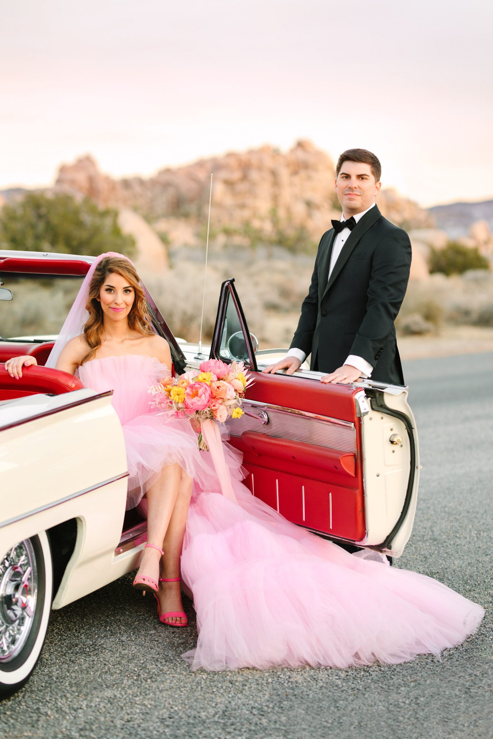 Bride and groom posing with white classic convertible car | Pink wedding dress Joshua Tree elopement featured on Green Wedding Shoes | Colorful desert wedding inspiration for fun-loving couples in Southern California #joshuatreewedding #joshuatreeelopement #pinkwedding #greenweddingshoes Source: Mary Costa Photography | Los Angeles