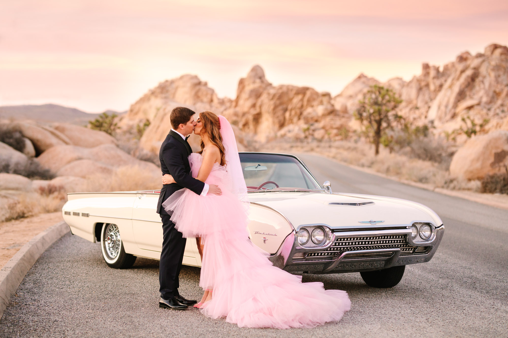 Bride and groom kissing in front of white classic convertible car | Pink wedding dress Joshua Tree elopement featured on Green Wedding Shoes | Colorful desert wedding inspiration for fun-loving couples in Southern California #joshuatreewedding #joshuatreeelopement #pinkwedding #greenweddingshoes Source: Mary Costa Photography | Los Angeles