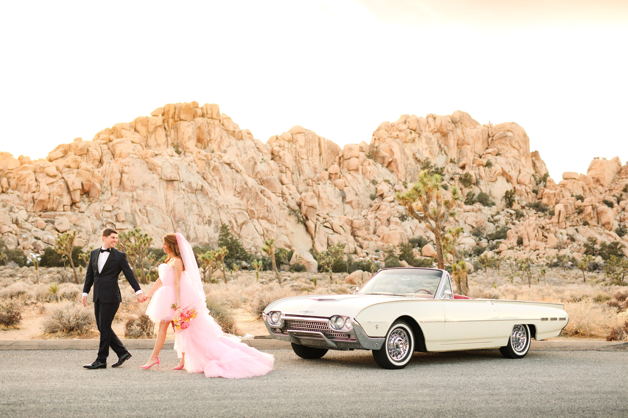 Bride and groom walking with white classic convertible | Pink wedding dress Joshua Tree elopement featured on Green Wedding Shoes | Colorful desert wedding inspiration for fun-loving couples in Southern California #joshuatreewedding #joshuatreeelopement #pinkwedding #greenweddingshoes Source: Mary Costa Photography | Los Angeles