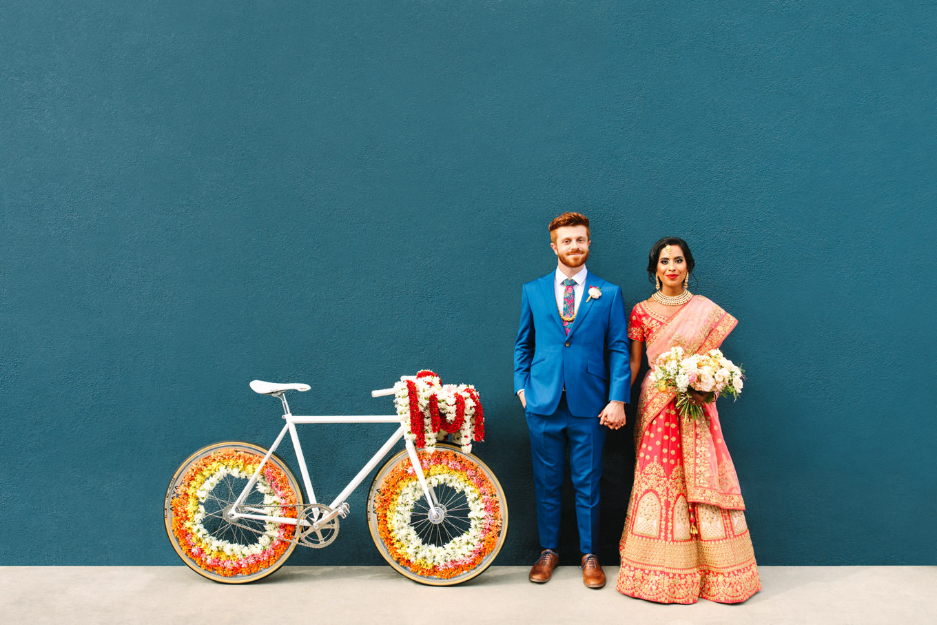Bride in red saree and groom in bright blue suit with floral Baraat bicycle on teal wall. Two Disney artists create a unique and colorful Indian Fusion wedding at The Fig House Los Angeles, featured on Green Wedding Shoes. | Colorful and elevated wedding inspiration for fun-loving couples in Southern California | #indianwedding #indianfusionwedding #thefighouse #losangeleswedding   Source: Mary Costa Photography | Los Angeles