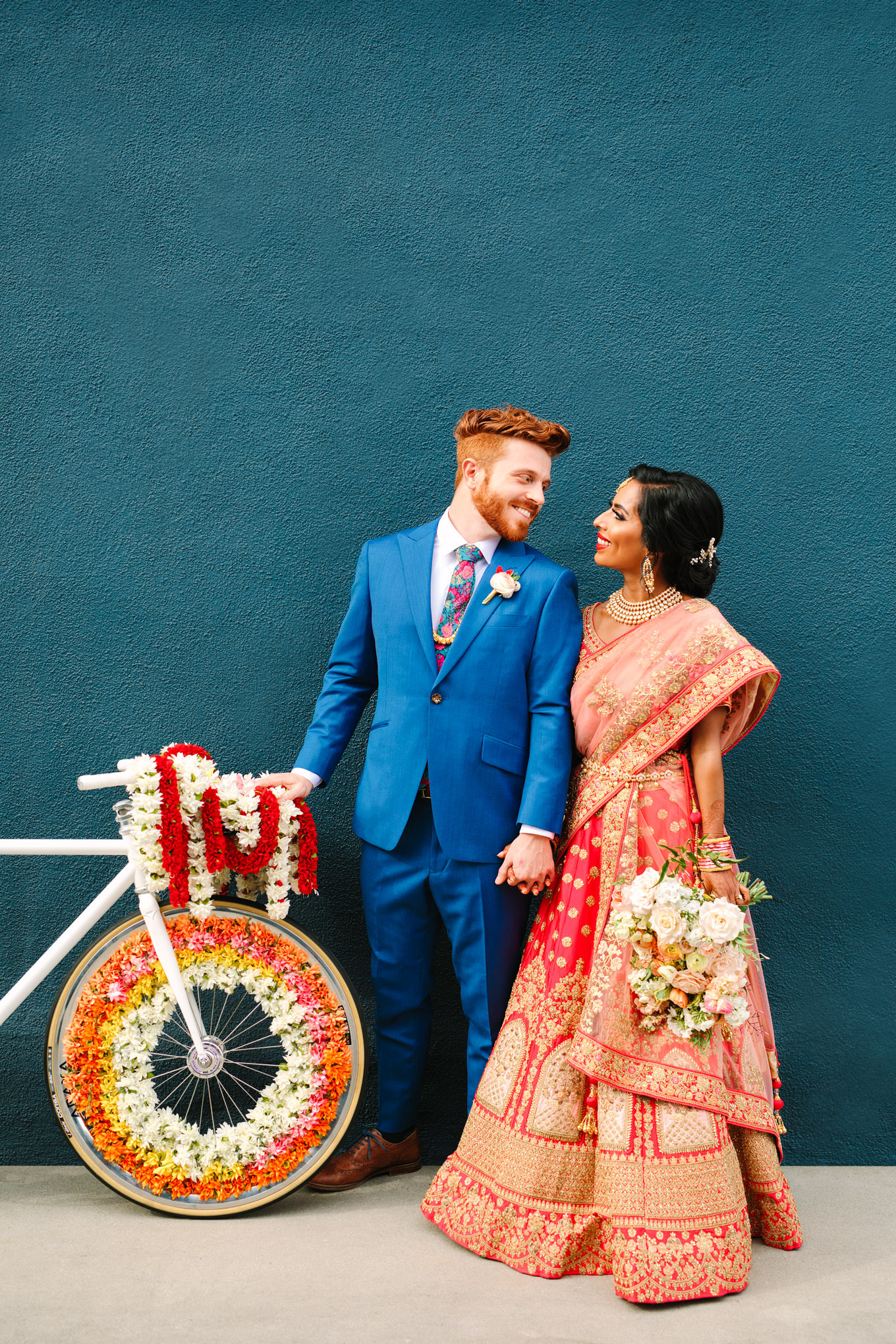 Bride in red saree and groom in bright blue suit with floral Baraat bicycle on teal wall. Two Disney artists create a unique and colorful Indian Fusion wedding at The Fig House Los Angeles, featured on Green Wedding Shoes. | Colorful and elevated wedding inspiration for fun-loving couples in Southern California | #indianwedding #indianfusionwedding #thefighouse #losangeleswedding   Source: Mary Costa Photography | Los Angeles