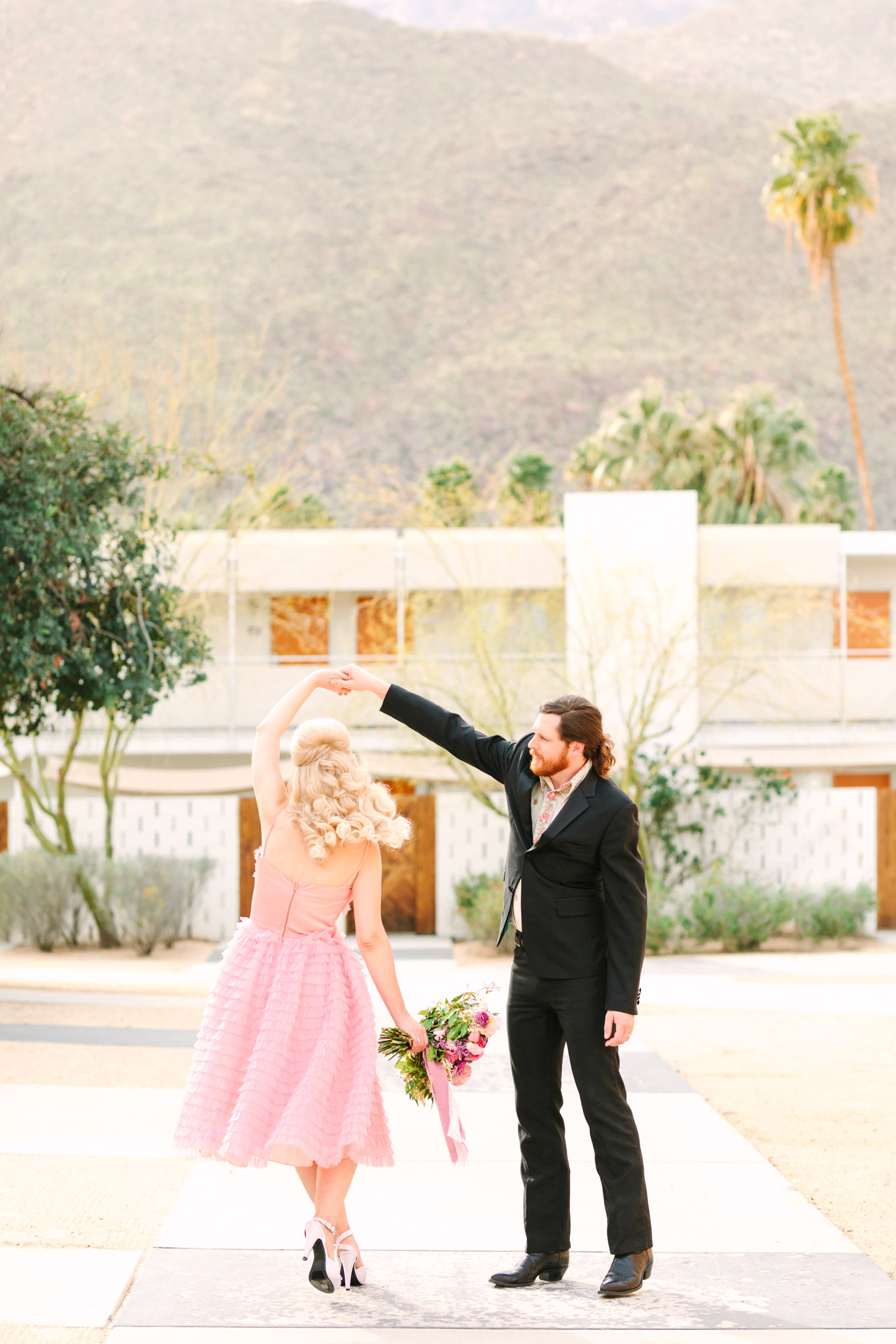 Couple twirling at Ace Hotel. Modern vintage-inspired Palm Springs engagement session with a 1960s pink Cadillac, retro clothing, and flowers by Shindig Chic. | Colorful and elevated wedding inspiration for fun-loving couples in Southern California | #engagementsession #PalmSpringsengagement #vintageweddingdress #floralcar #pinkcar   Source: Mary Costa Photography | Los Angeles