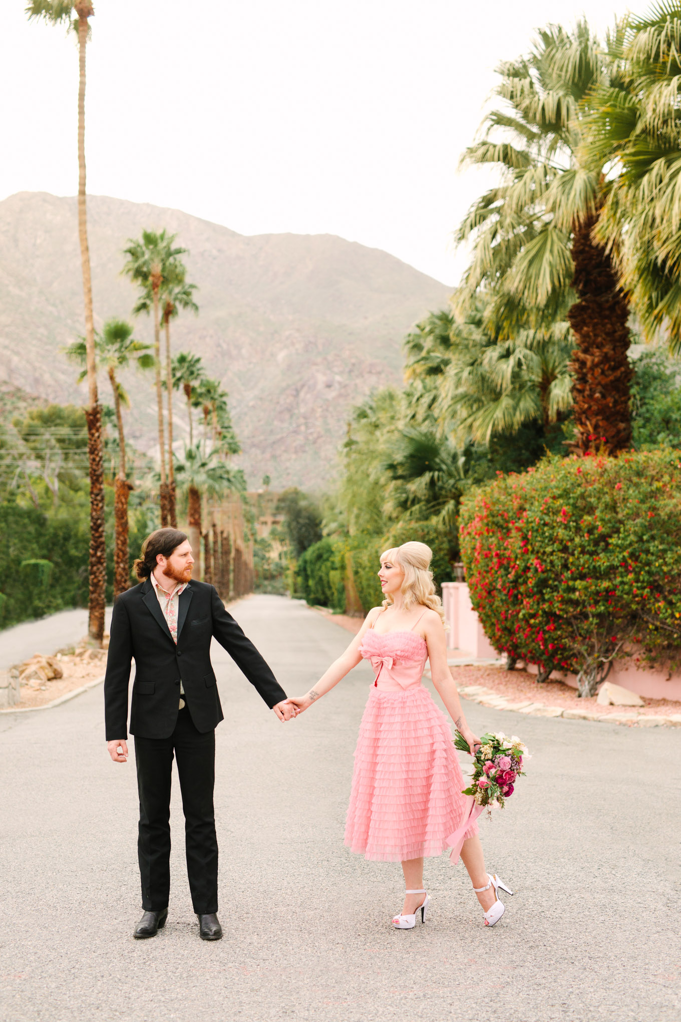 Couple walking among palm trees. Modern vintage-inspired Palm Springs engagement session with a 1960s pink Cadillac, retro clothing, and flowers by Shindig Chic. | Colorful and elevated wedding inspiration for fun-loving couples in Southern California | #engagementsession #PalmSpringsengagement #vintageweddingdress #floralcar #pinkcar   Source: Mary Costa Photography | Los Angeles