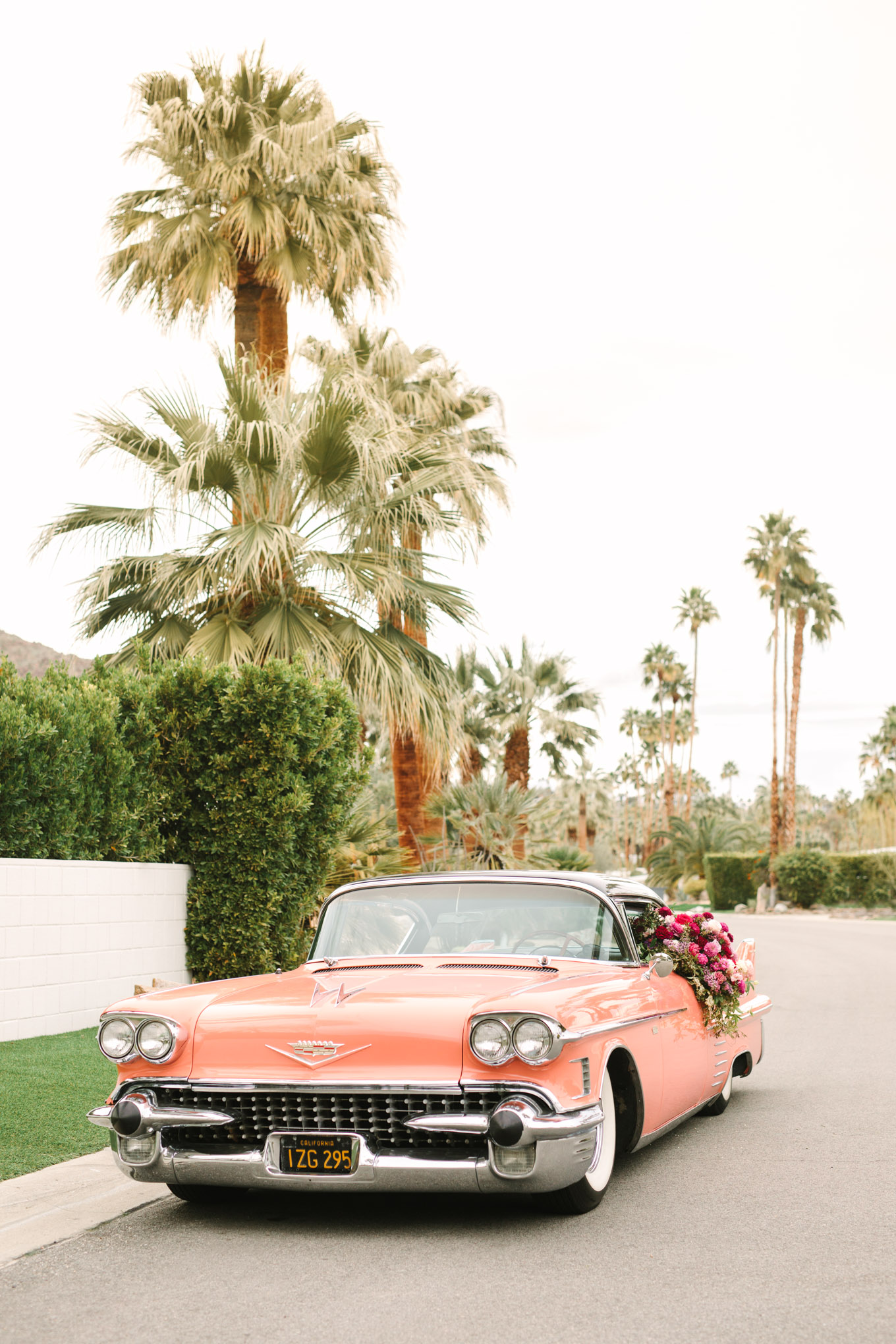 Classic car parked in Mid Century Modern neighborhood. Modern vintage-inspired Palm Springs engagement session with a 1960s pink Cadillac, retro clothing, and flowers by Shindig Chic. | Colorful and elevated wedding inspiration for fun-loving couples in Southern California | #engagementsession #PalmSpringsengagement #vintageweddingdress #floralcar #pinkcar   Source: Mary Costa Photography | Los Angeles