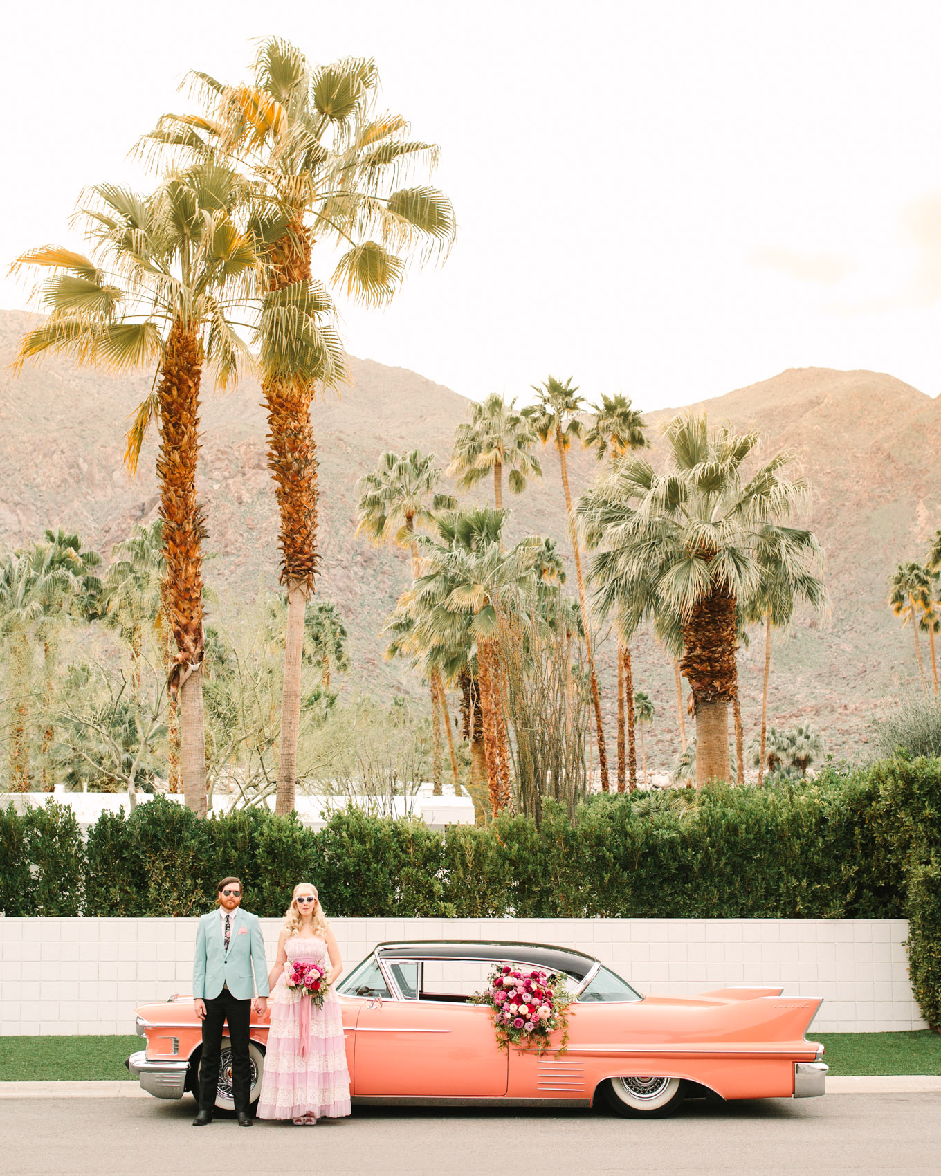 Couple with pink classic car. Modern vintage-inspired Palm Springs engagement session with a 1960s Cadillac, retro clothing, and flowers by Shindig Chic. | Colorful and elevated wedding inspiration for fun-loving couples in Southern California | #engagementsession #PalmSpringsengagement #vintageweddingdress #floralcar #pinkcar   Source: Mary Costa Photography | Los Angeles