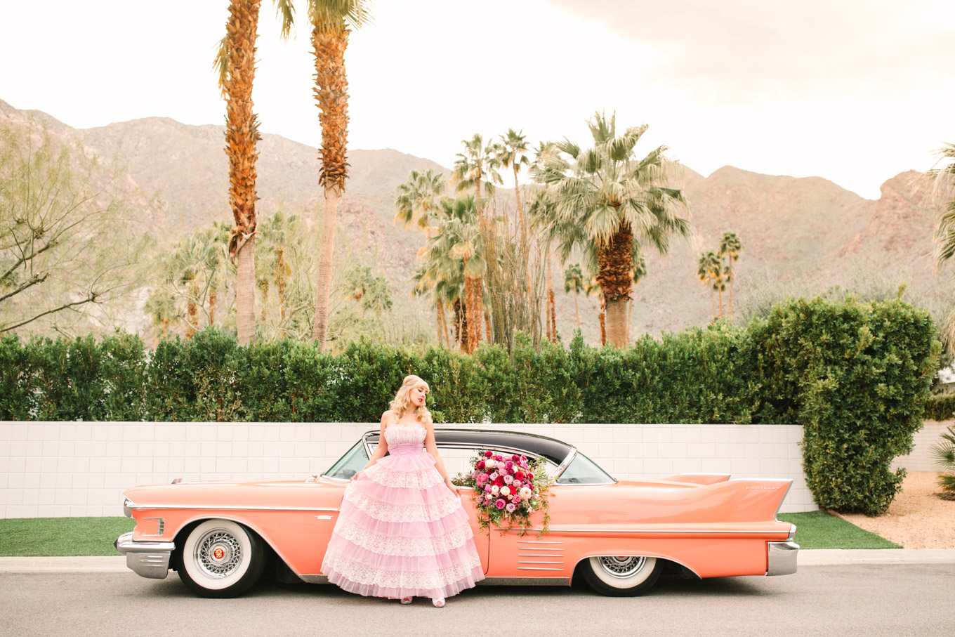 Bride with coral classic car. Modern vintage-inspired Palm Springs engagement session with a 1960s pink Cadillac, retro clothing, and flowers by Shindig Chic. | Colorful and elevated wedding inspiration for fun-loving couples in Southern California | #engagementsession #PalmSpringsengagement #vintageweddingdress #floralcar #pinkcar   Source: Mary Costa Photography | Los Angeles