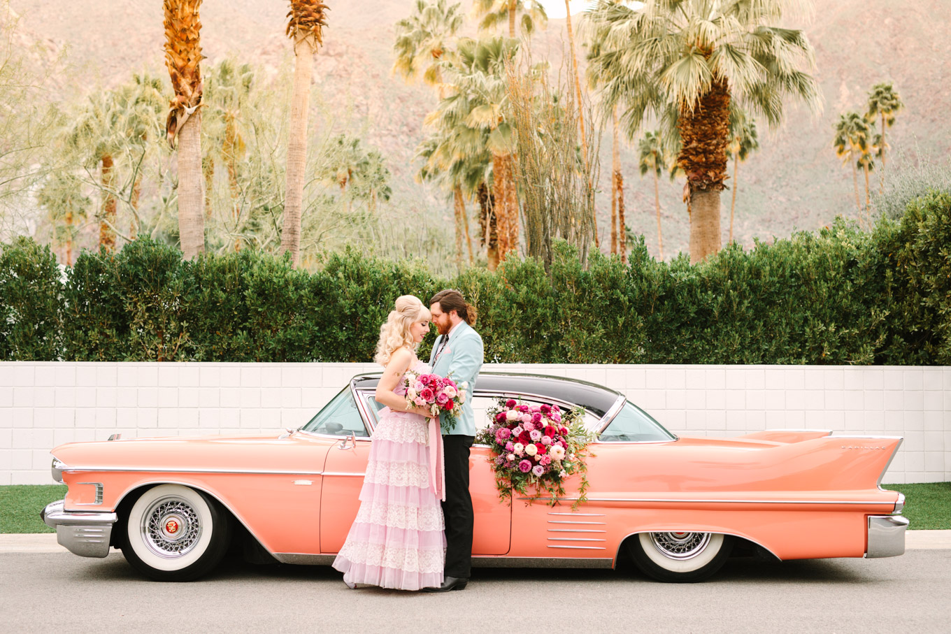 Couple embracing in front of pink classic car. Modern vintage-inspired Palm Springs engagement session with a 1960s pink Cadillac, retro clothing, and flowers by Shindig Chic. | Colorful and elevated wedding inspiration for fun-loving couples in Southern California | #engagementsession #PalmSpringsengagement #vintageweddingdress #floralcar #pinkcar   Source: Mary Costa Photography | Los Angeles