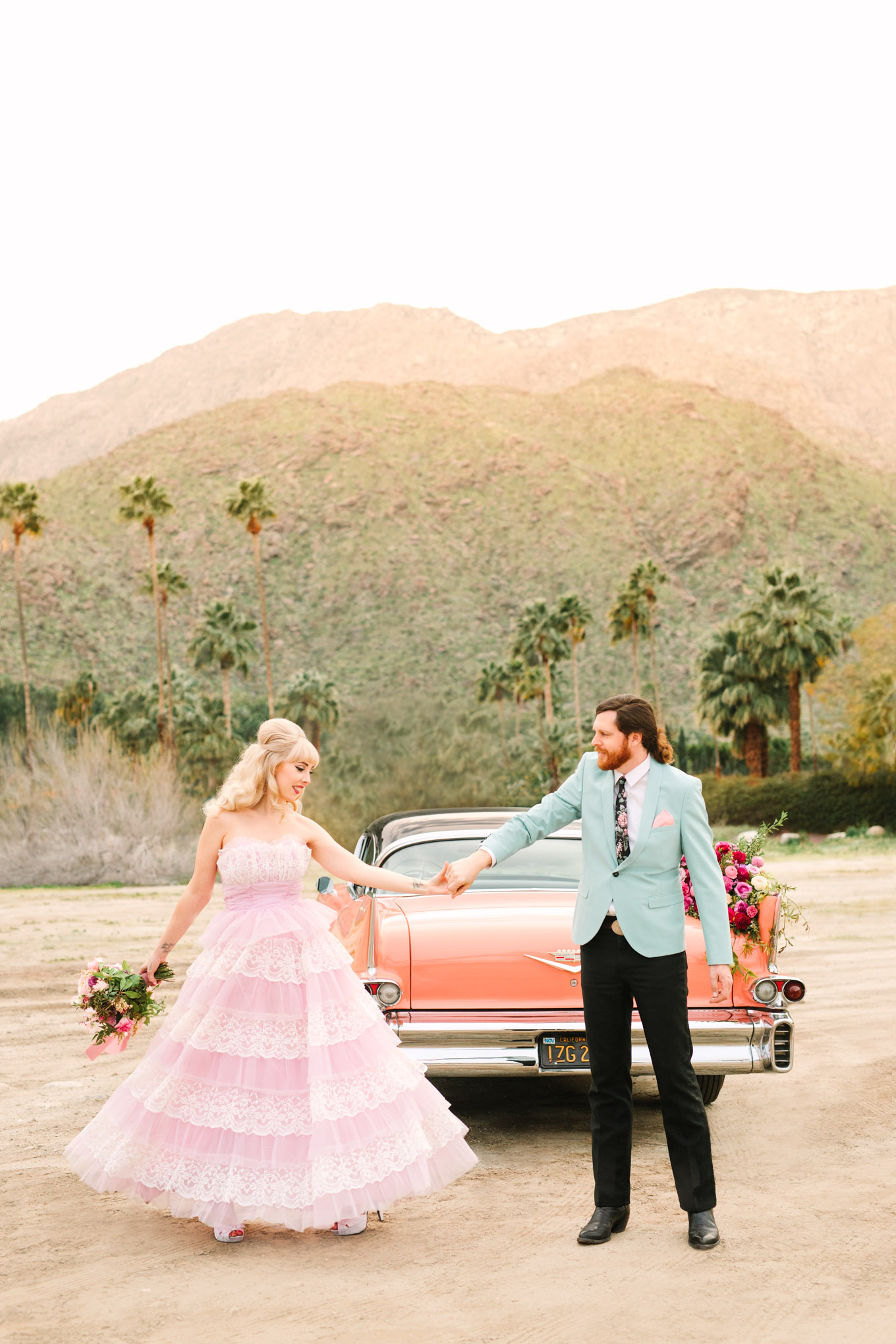 Bride and groom twirling with classic car. Modern vintage-inspired Palm Springs engagement session with a 1960s pink Cadillac, retro clothing, and flowers by Shindig Chic. | Colorful and elevated wedding inspiration for fun-loving couples in Southern California | #engagementsession #PalmSpringsengagement #vintageweddingdress #floralcar #pinkcar   Source: Mary Costa Photography | Los Angeles