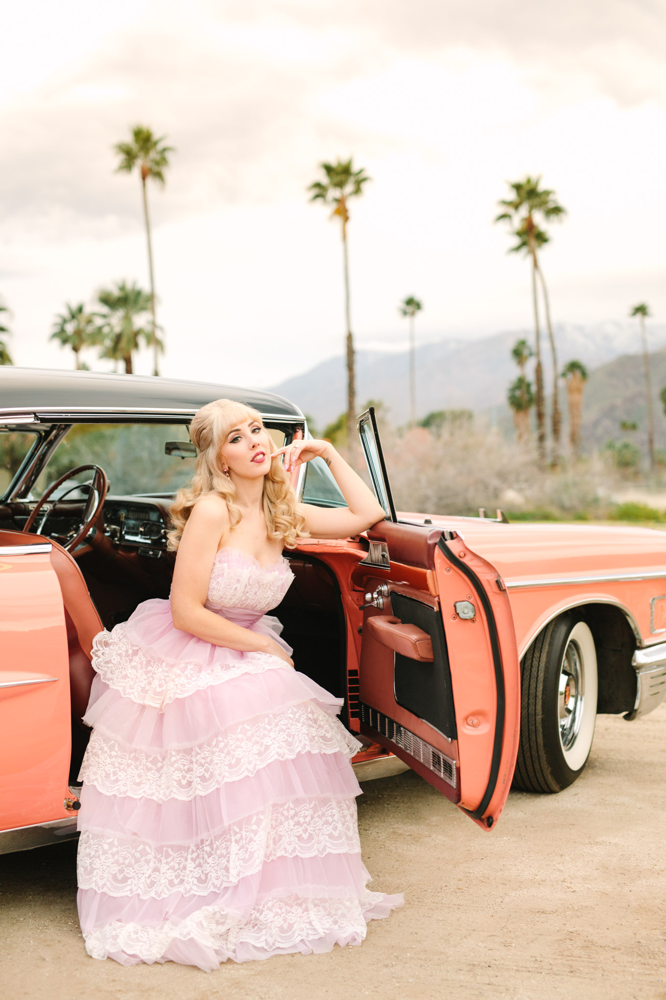 Bride sitting in classic car. Modern vintage-inspired Palm Springs engagement session with a 1960s pink Cadillac, retro clothing, and flowers by Shindig Chic. | Colorful and elevated wedding inspiration for fun-loving couples in Southern California | #engagementsession #PalmSpringsengagement #vintageweddingdress #floralcar #pinkcar   Source: Mary Costa Photography | Los Angeles