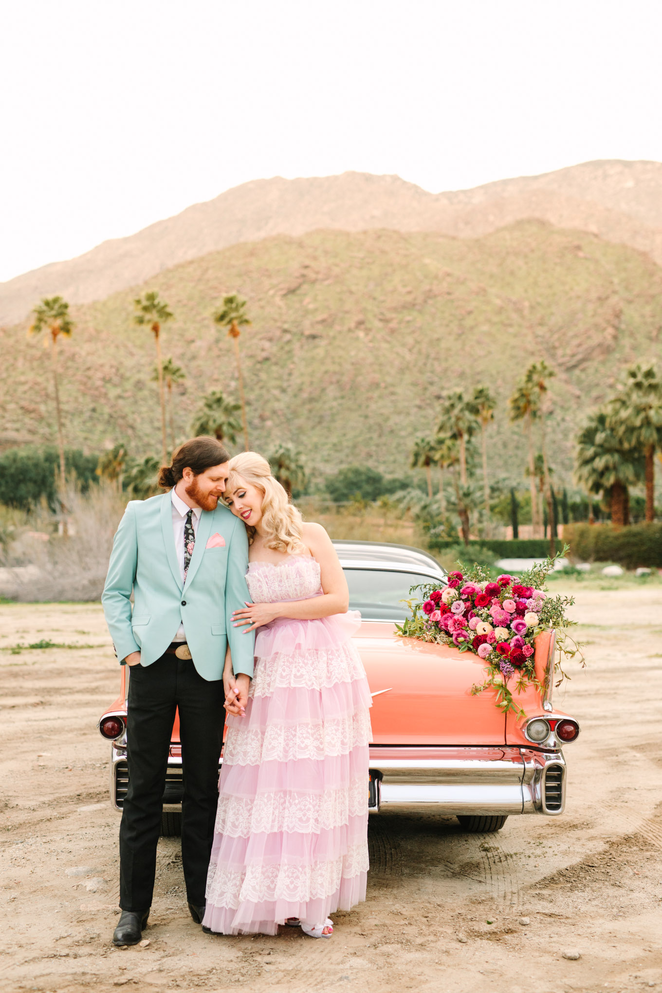 Bride and groom with classic car. Modern vintage-inspired Palm Springs engagement session with a 1960s pink Cadillac, retro clothing, and flowers by Shindig Chic. | Colorful and elevated wedding inspiration for fun-loving couples in Southern California | #engagementsession #PalmSpringsengagement #vintageweddingdress #floralcar #pinkcar   Source: Mary Costa Photography | Los Angeles