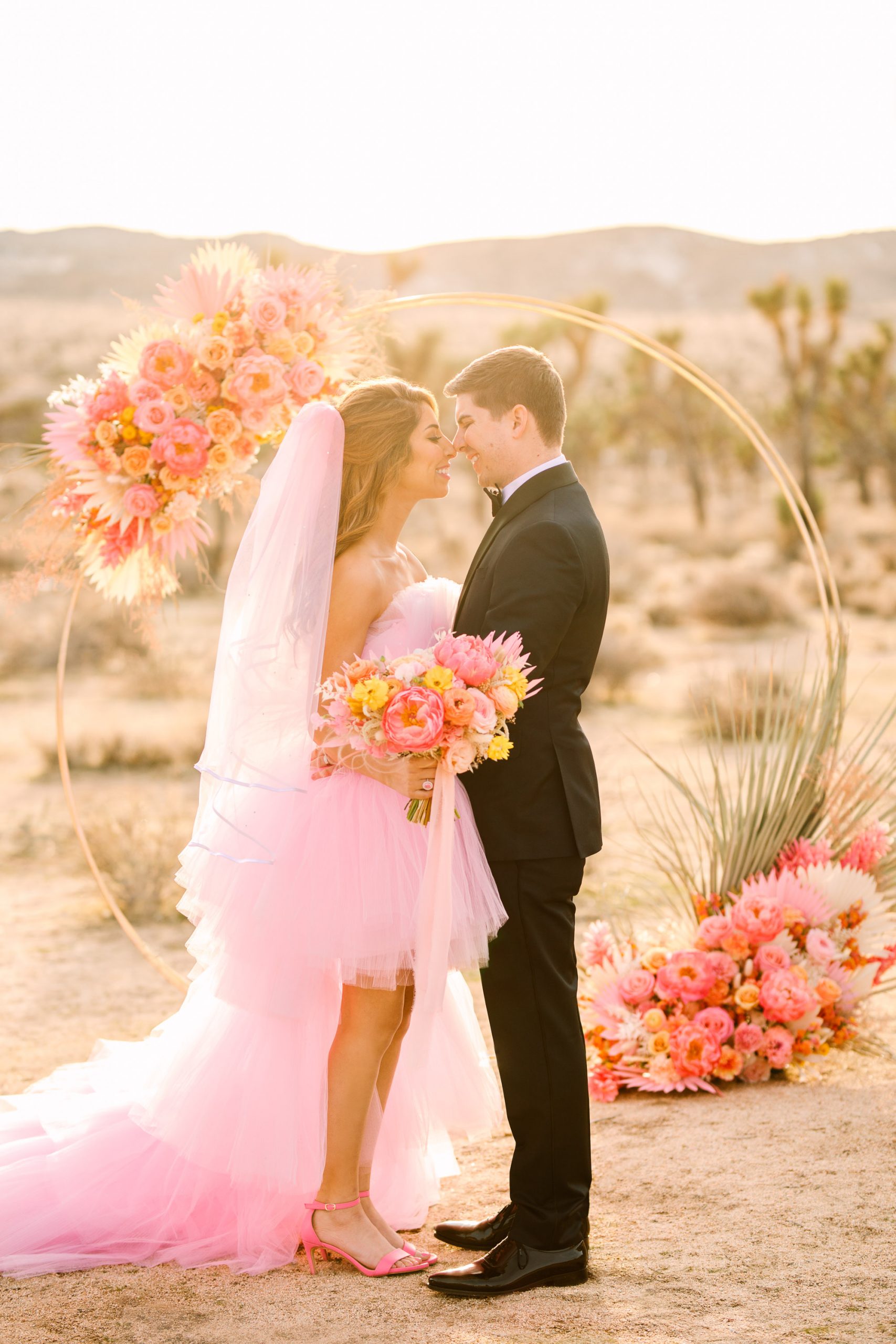 Bride and groom in front of circular ceremony arch | Pink wedding dress Joshua Tree elopement featured on Green Wedding Shoes | Colorful desert wedding inspiration for fun-loving couples in Southern California #joshuatreewedding #joshuatreeelopement #pinkwedding #greenweddingshoes Source: Mary Costa Photography | Los Angeles