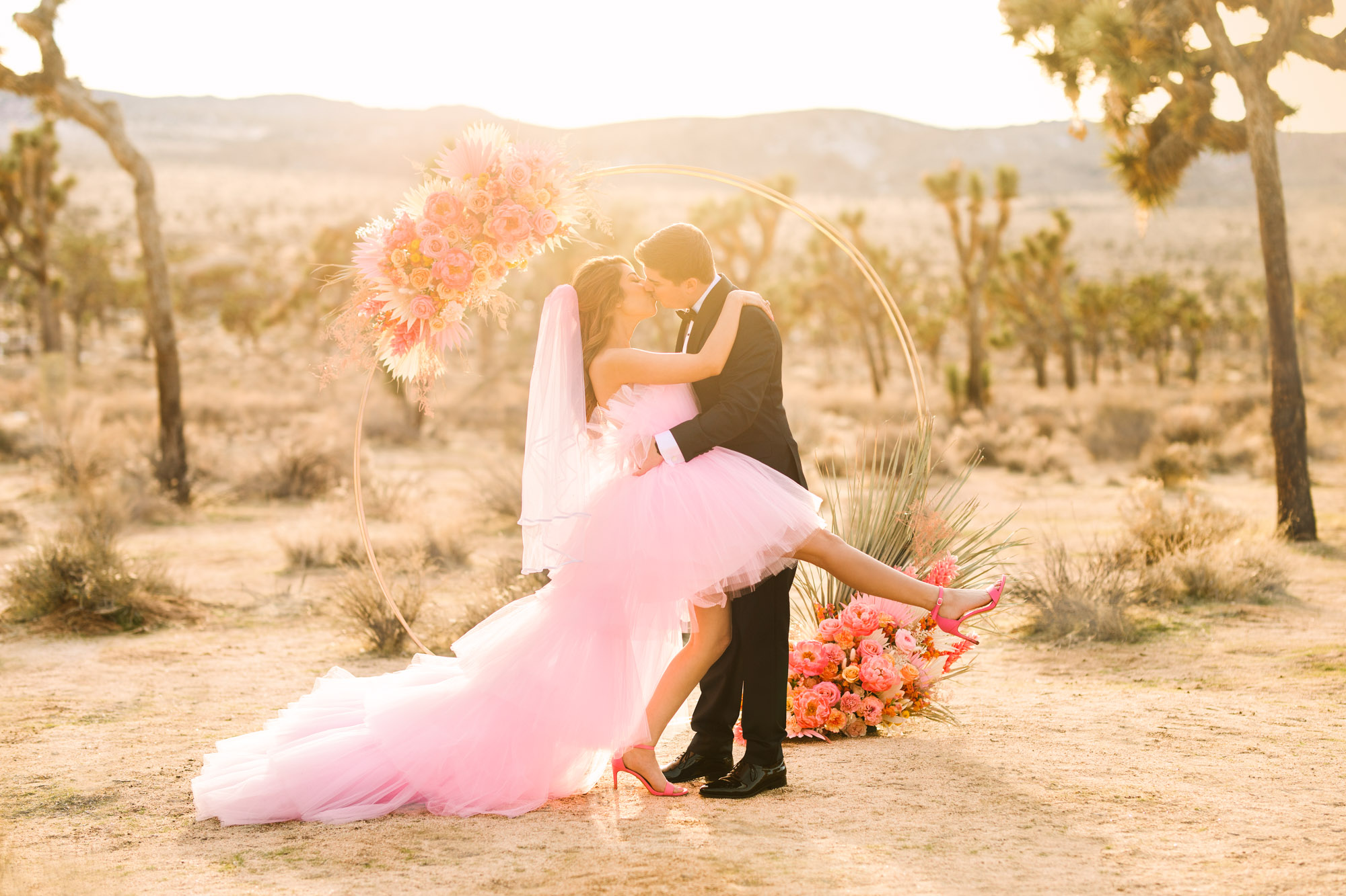 Bride and groom kissing in front of floral circular ceremony arch | Pink wedding dress Joshua Tree elopement featured on Green Wedding Shoes | Colorful desert wedding inspiration for fun-loving couples in Southern California #joshuatreewedding #joshuatreeelopement #pinkwedding #greenweddingshoes Source: Mary Costa Photography | Los Angeles