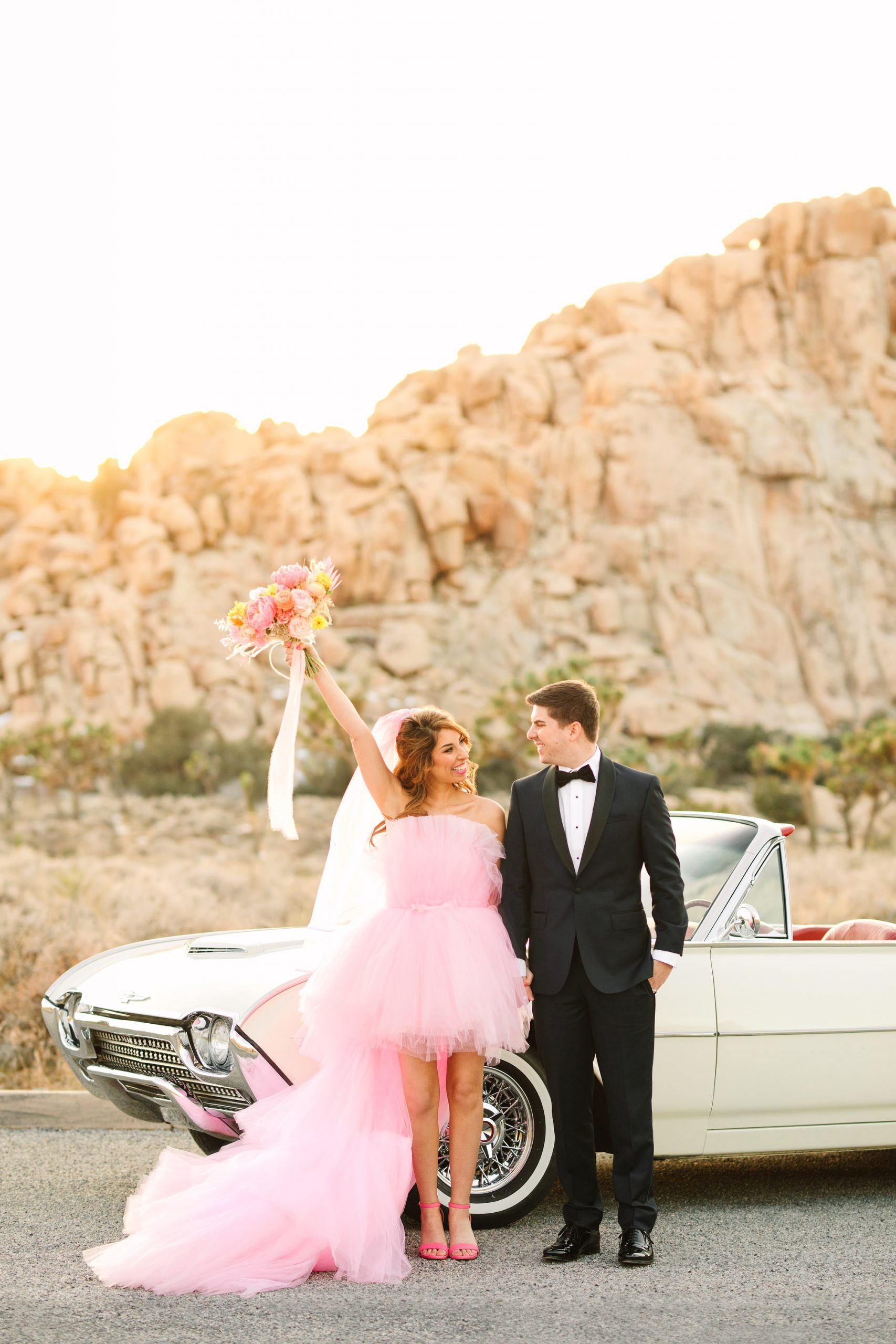 Bride and groom with white classic Ford Thunderbird convertible | Pink wedding dress Joshua Tree elopement featured on Green Wedding Shoes | Colorful desert wedding inspiration for fun-loving couples in Southern California #joshuatreewedding #joshuatreeelopement #pinkwedding #greenweddingshoes Source: Mary Costa Photography | Los Angeles