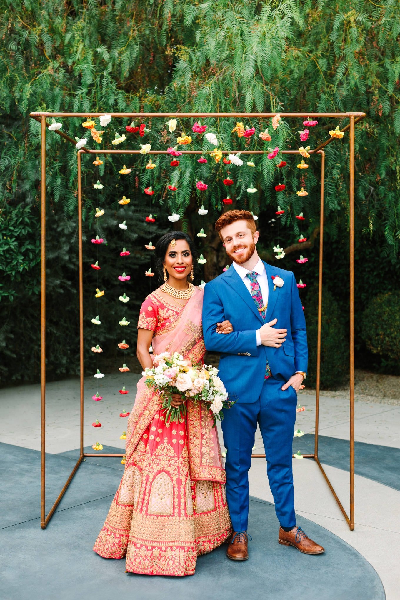 Bride in red saree and groom in bright blue suit in front of carnation wedding ceremony arch. Two Disney artists create a unique and colorful Indian Fusion wedding at The Fig House Los Angeles, featured on Green Wedding Shoes. | Colorful and elevated wedding inspiration for fun-loving couples in Southern California | #indianwedding #indianfusionwedding #thefighouse #losangeleswedding   Source: Mary Costa Photography | Los Angeles