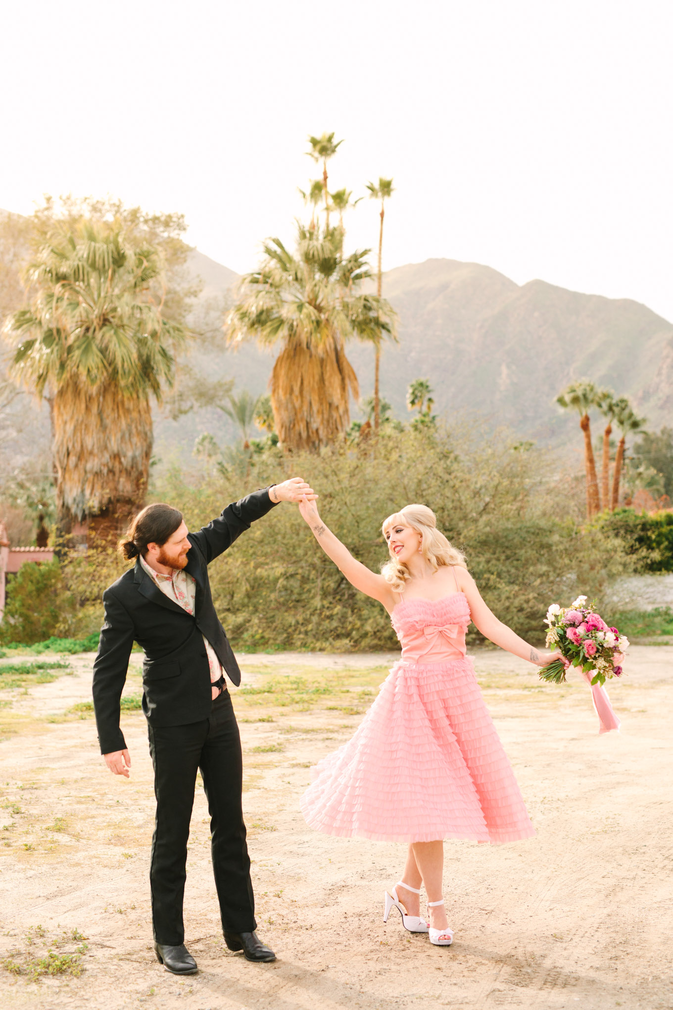 Retro couple twirling among palm trees. Modern vintage-inspired Palm Springs engagement session with a 1960s pink Cadillac, retro clothing, and flowers by Shindig Chic. | Colorful and elevated wedding inspiration for fun-loving couples in Southern California | #engagementsession #PalmSpringsengagement #vintageweddingdress #floralcar #pinkcar   Source: Mary Costa Photography | Los Angeles